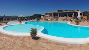 A large outdoor swimming pool with a curved design, surrounded by a paved area with potted plants. - Parco Degli Ulivi (Arzachena)