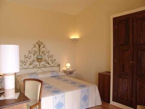 A bedroom with a bed, a chair, a nightstand with a lamp, and a wooden wardrobe. - Parco Degli Ulivi (Arzachena)