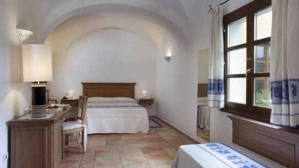 A bedroom with a bed, bedside tables, a desk, a lamp, a chair, and a window with curtains. - Parco Degli Ulivi (Arzachena)