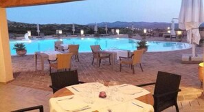 An outdoor pool area with chairs and tables, a view of mountains, and a dining setup. - Parco Degli Ulivi (Arzachena)