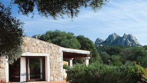 A stone building with a covered outdoor seating area, surrounded by greenery and a mountainous backdrop. - Parco Degli Ulivi (Arzachena)