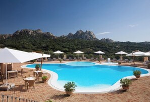 A swimming pool with lounge chairs and umbrellas, surrounded by brick flooring and potted plants. - Parco Degli Ulivi (Arzachena)