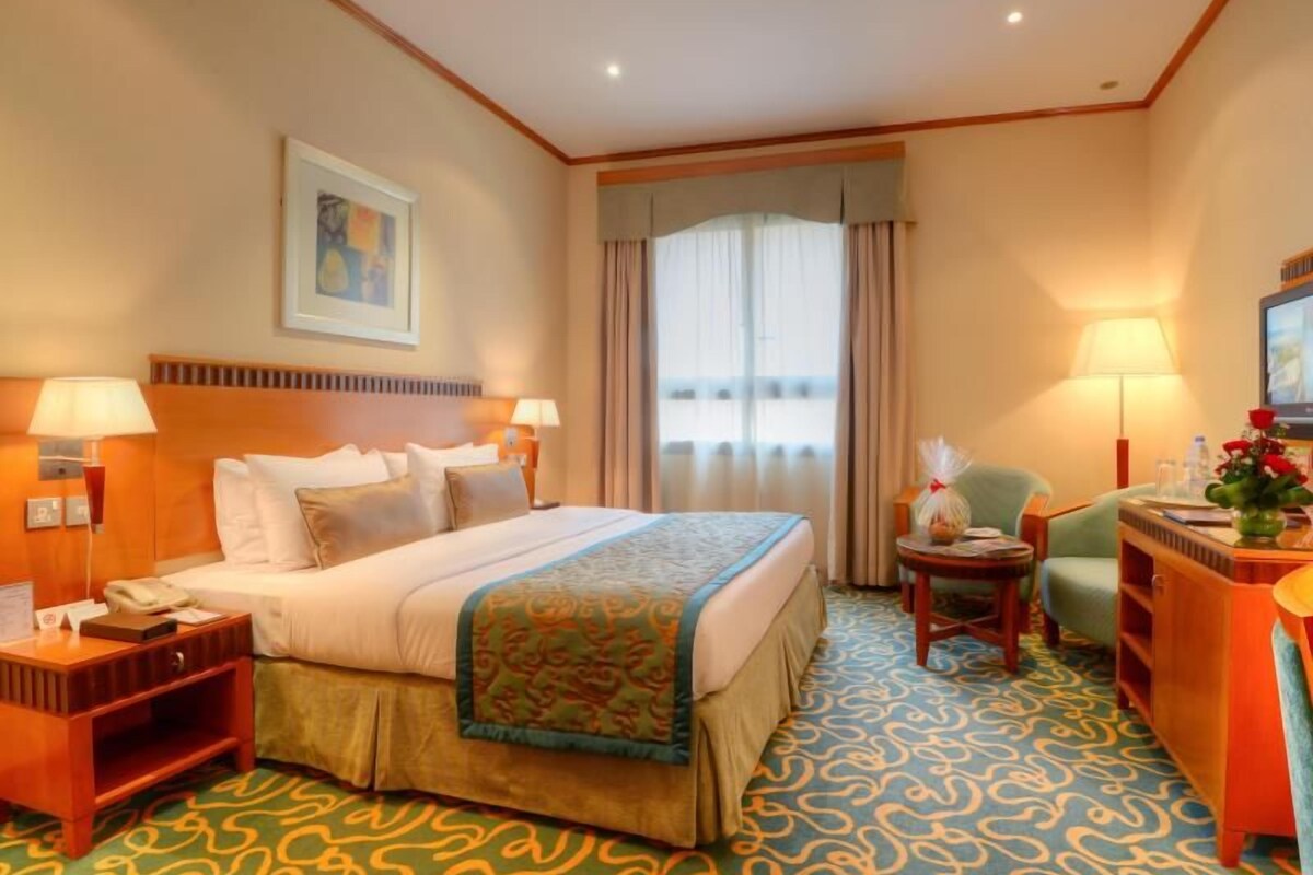 Ein Hotelzimmer mit einem großen Bett, zwei Nachttischen, einem Schreibtisch mit Stuhl, einem Fernseher und einem Fenster mit Vorhängen.