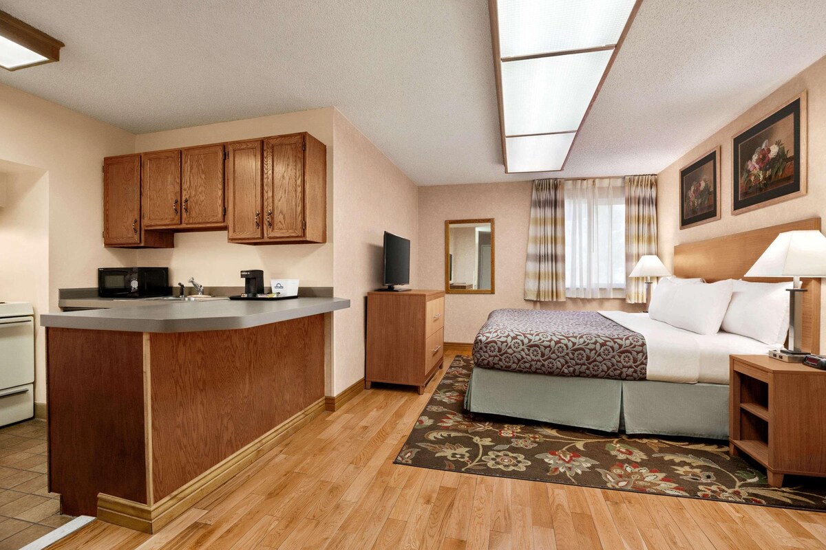 A hotel room with a kitchenette, a bed with a patterned bedspread, a bedside table with a lamp, a large mirror, and a floral rug.