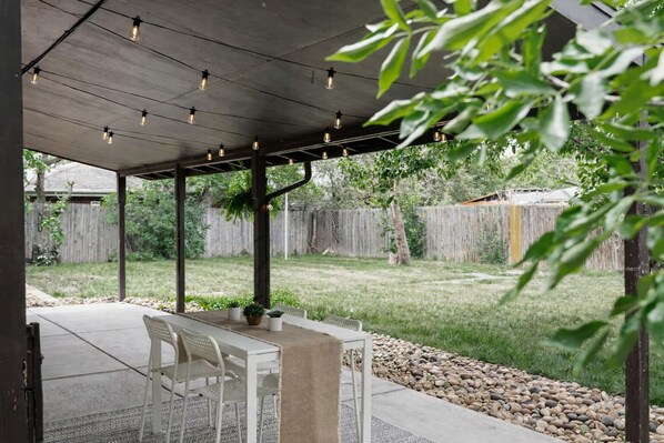 Un patio coperto con tavolo e sedie, un pergolato con luci a stringa e un giardino sul retro con recinzione in legno e vegetazione.