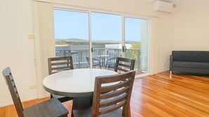 A room with a round table, four chairs, a sofa, and a sliding glass door leading to a balcony with a view of hills. - Loftberg Villas (Muswellbrook)