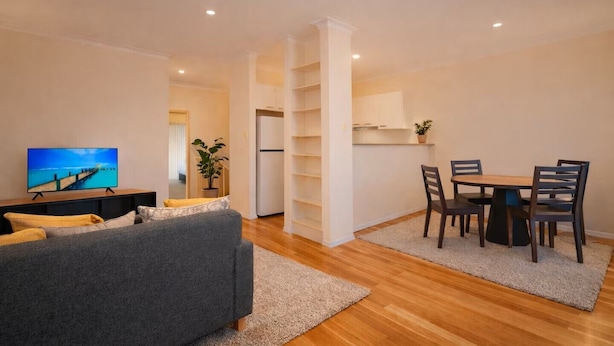 A living room with a TV, sofa, dining table, chairs, and a kitchen area. - Loftberg Villas (Muswellbrook)