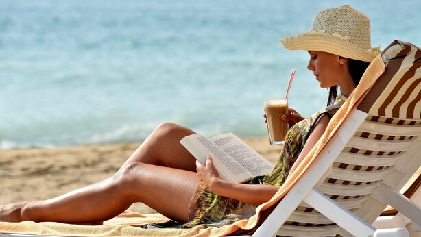 A person reclining on a beach chair, reading a book, with a drink in hand.
