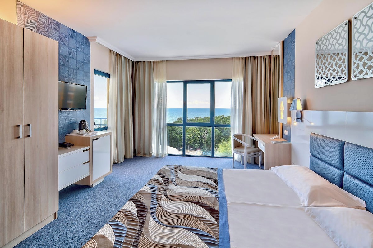 Ein Hotelzimmer mit einem großen Bett, einem Schreibtisch, einem Fernseher und Meerblick.