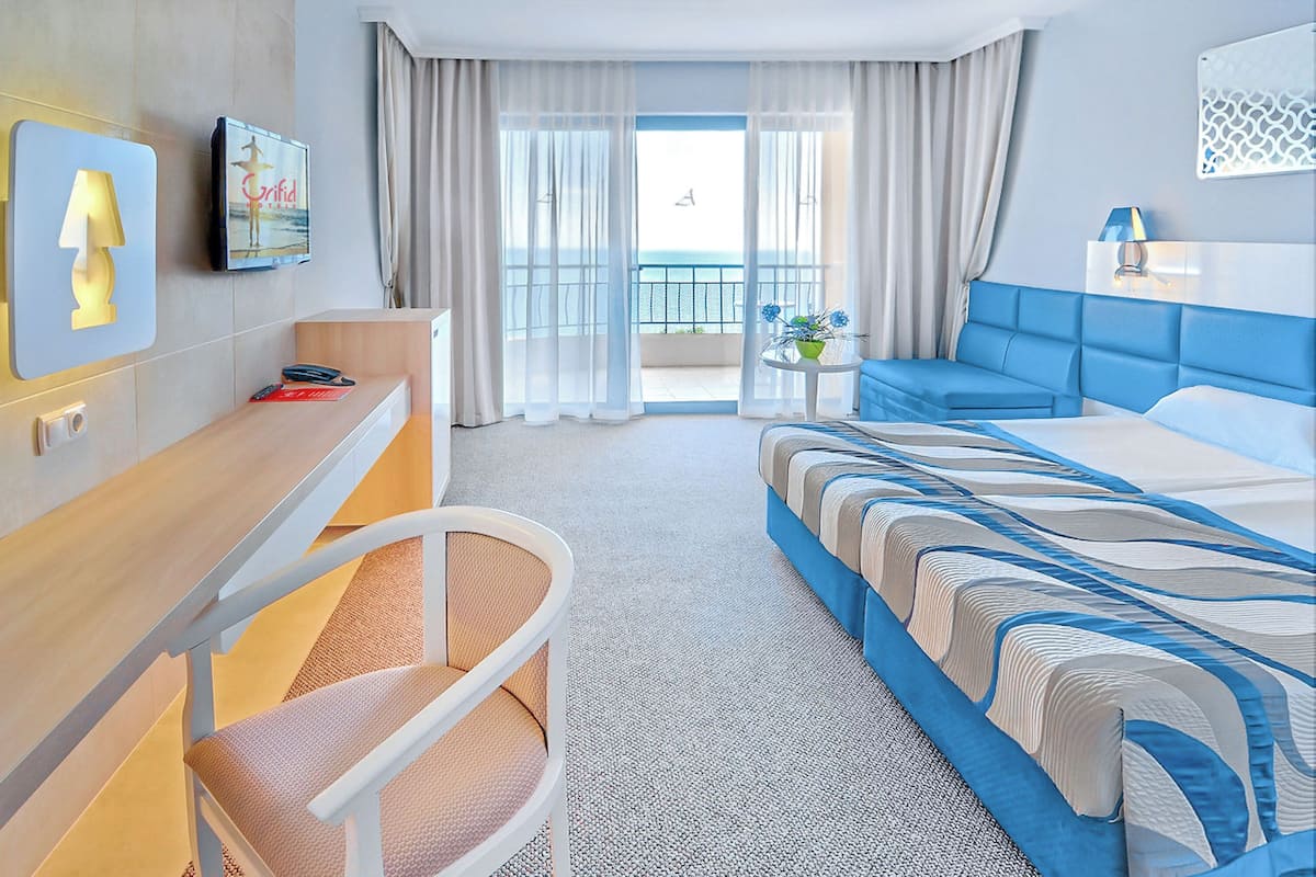 Ein Hotelzimmer mit einem Bett, einem Schreibtisch, einem Sessel, einem kleinen Tisch mit einer Vase und Meerblick durch große Fenster.