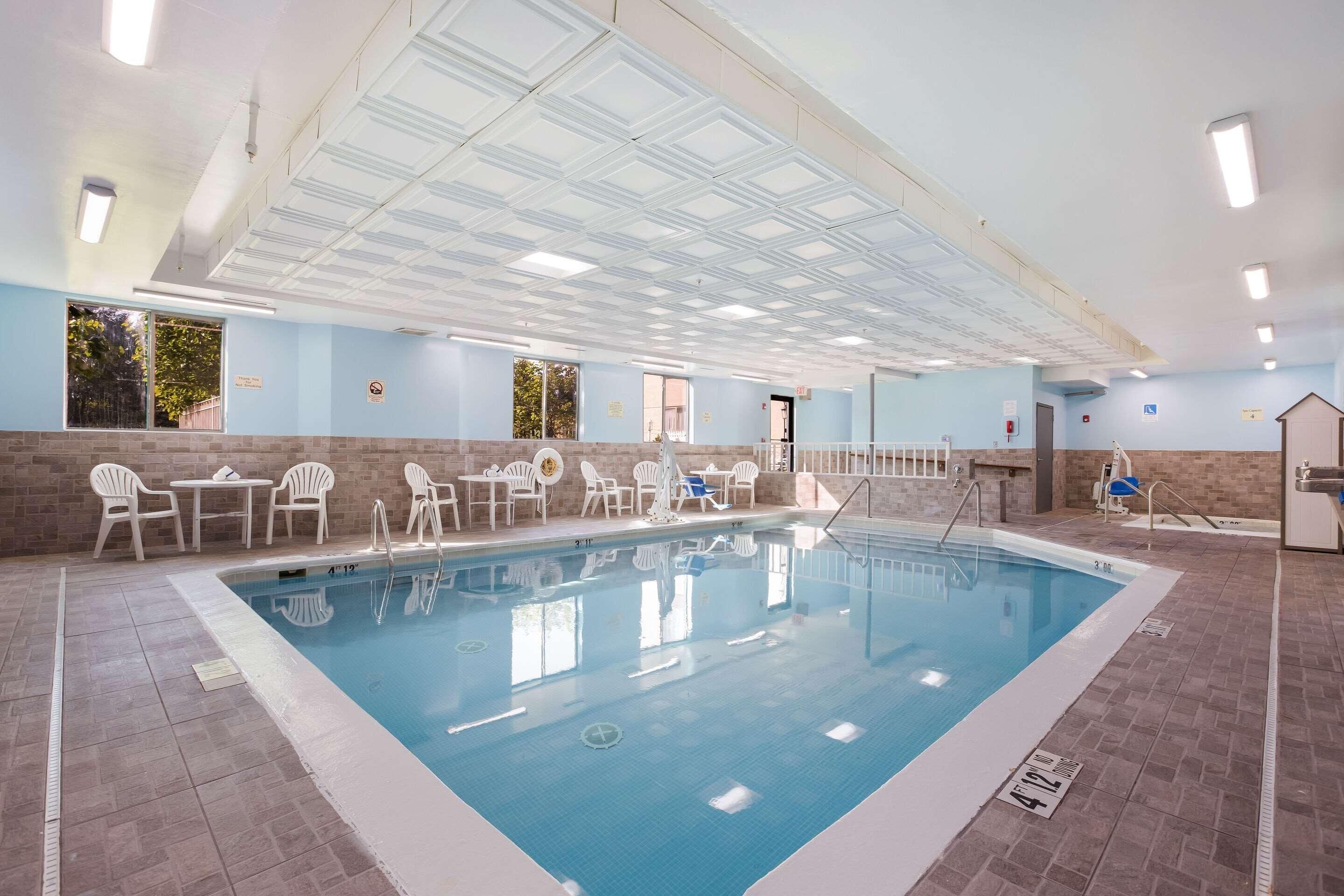 An indoor swimming pool with clear water, surrounded by white chairs and tables, and a tiled floor.