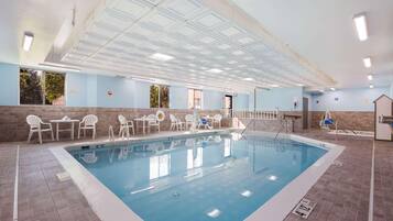 An indoor swimming pool with clear water, surrounded by white chairs and tables, and a tiled floor.