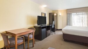 A hotel room with a bed, a desk with a television, a chair, and a small refrigerator.