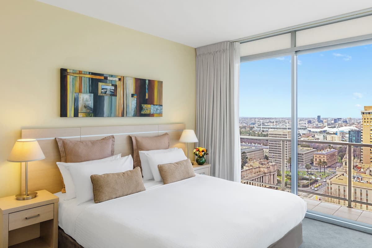 Habitación de hotel con una cama grande, una mesita de noche con lámpara, un jarrón con flores y vistas a la ciudad a través de amplios ventanales.