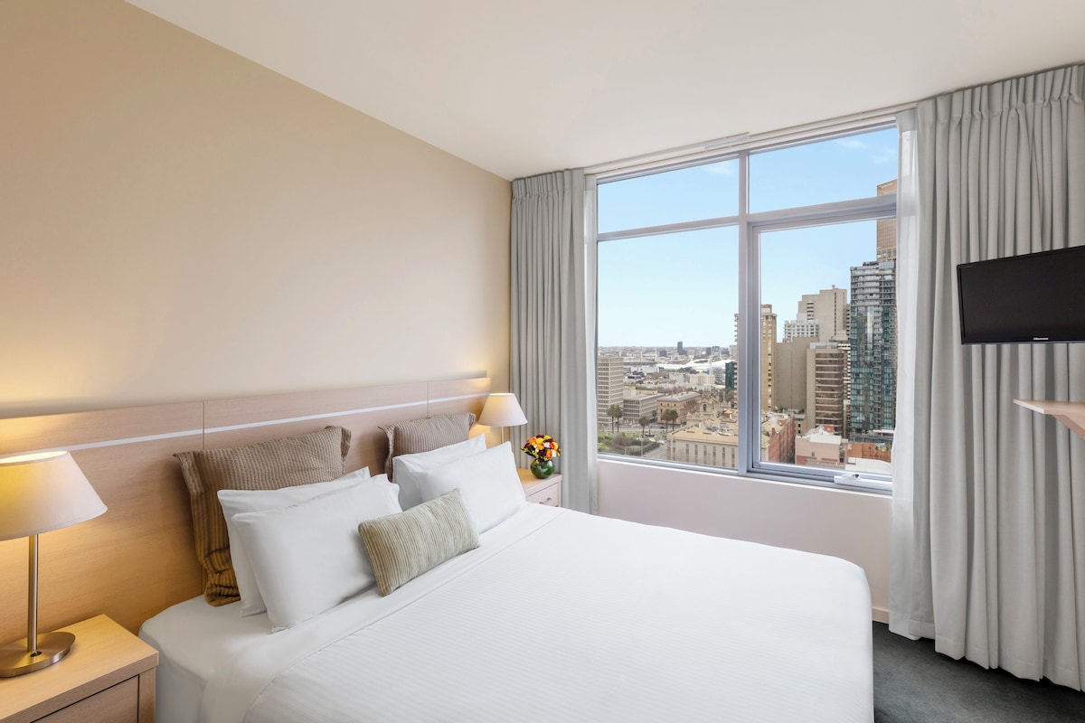 A hotel room with a large bed, a nightstand with a lamp, a TV on a stand, and a window with a city view.