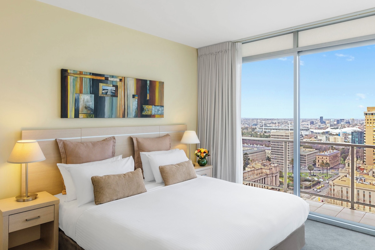 A hotel room with a large bed, a nightstand with a lamp, a vase with flowers, and a city view through large windows.