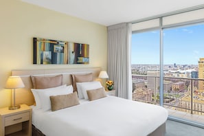 A hotel room with a large bed, a nightstand with a lamp, a vase with flowers, and a city view through large windows. - Oaks Melbourne Lonsdale Suites (Melbourne)
