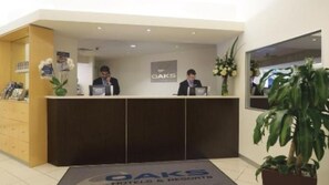 A reception desk with two staff members, a logo for OAKS Hotels & Resorts, and a reception area with a plant and a mirror. - Oaks Melbourne Lonsdale Suites (Melbourne)