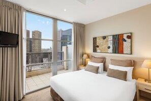 A hotel room with a large bed, two bedside tables with lamps, a flat-screen TV mounted on the wall, and a large window offering a city view. - Oaks Melbourne Lonsdale Suites (Melbourne)