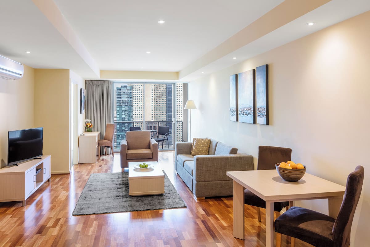Una sala moderna con un televisor de pantalla plana, un sofá, una mesa de comedor con un frutero, y vistas a la ciudad a través de amplios ventanales.