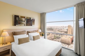A hotel room with a large bed, a nightstand, a lamp, a TV, and a city view through large windows. - Oaks Melbourne Lonsdale Suites (Melbourne)