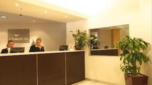 A reception desk with a sign that reads 'OAKS,' two receptionists, and potted plants. - Oaks Melbourne Lonsdale Suites (Melbourne)