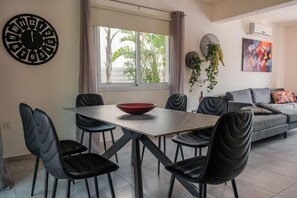 Ein Essbereich mit Tisch und Stühlen, einer Wanduhr, einem Fenster mit Vorhängen und einer Couch im Hintergrund.