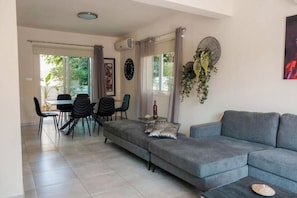Ein modernes Wohnzimmer mit einem grauen Sofa, einem Essbereich mit Tisch und Stühlen sowie einem großen Fenster mit Vorhängen.