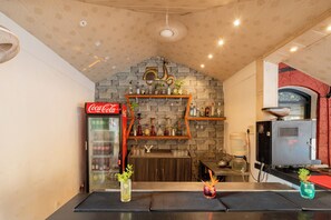 A bar area with a Coca-Cola refrigerator, shelves with bottles, a coffee machine, and a stone wall. - Kyriad Prestige Calangute Goa by Othpl (Calangute)