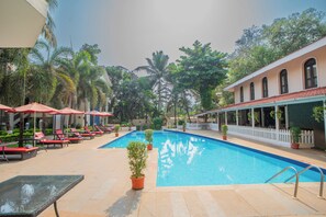 Una piscina con sillas de descanso y sombrillas, rodeada de vegetación y un edificio con valla blanca.