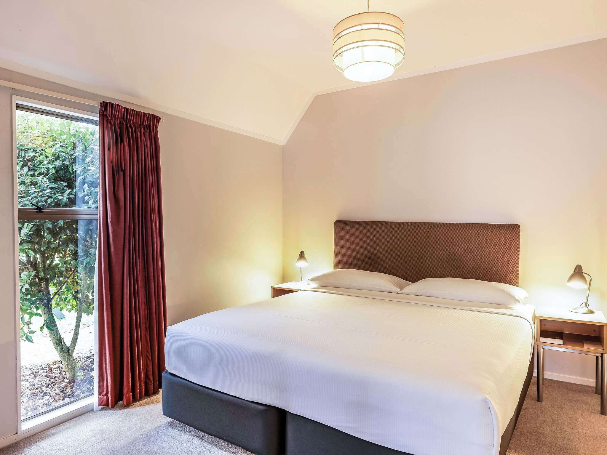 Ein Schlafzimmer mit Bett, Nachttisch, Lampe, Fenster mit Vorhängen und Blick auf Bäume.