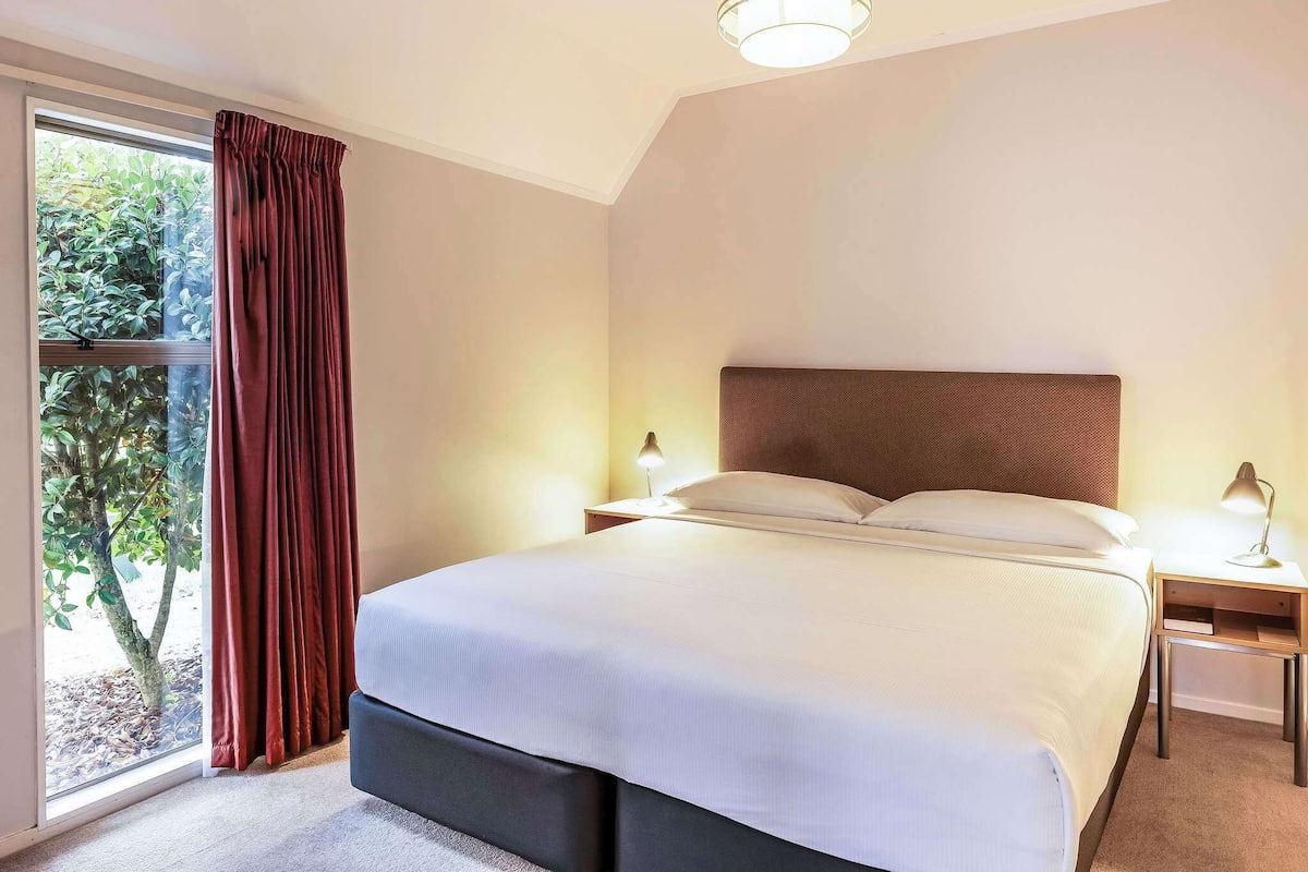 Un dormitorio con una cama, una mesita de noche, una lámpara, una ventana con cortinas y vistas a árboles.