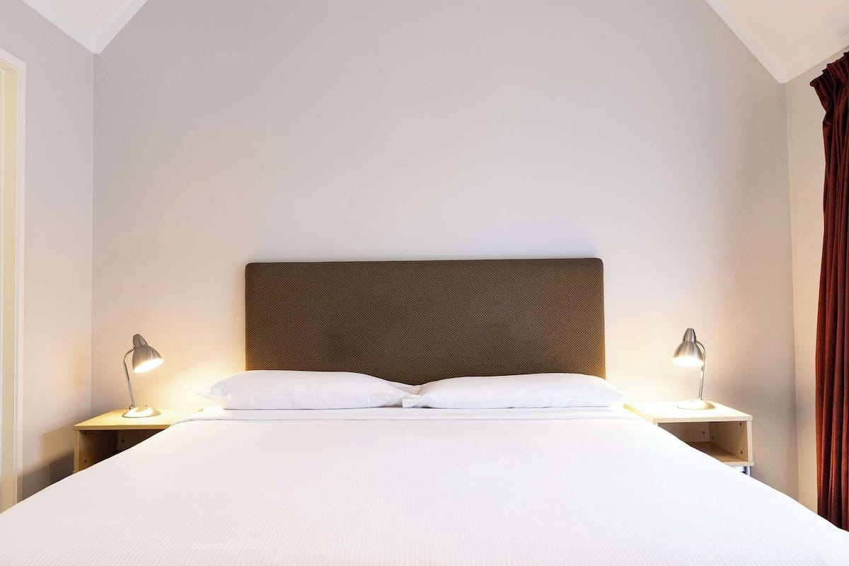 Una cama bien hecha con un cabecero, dos lámparas de noche y una mesita de madera.