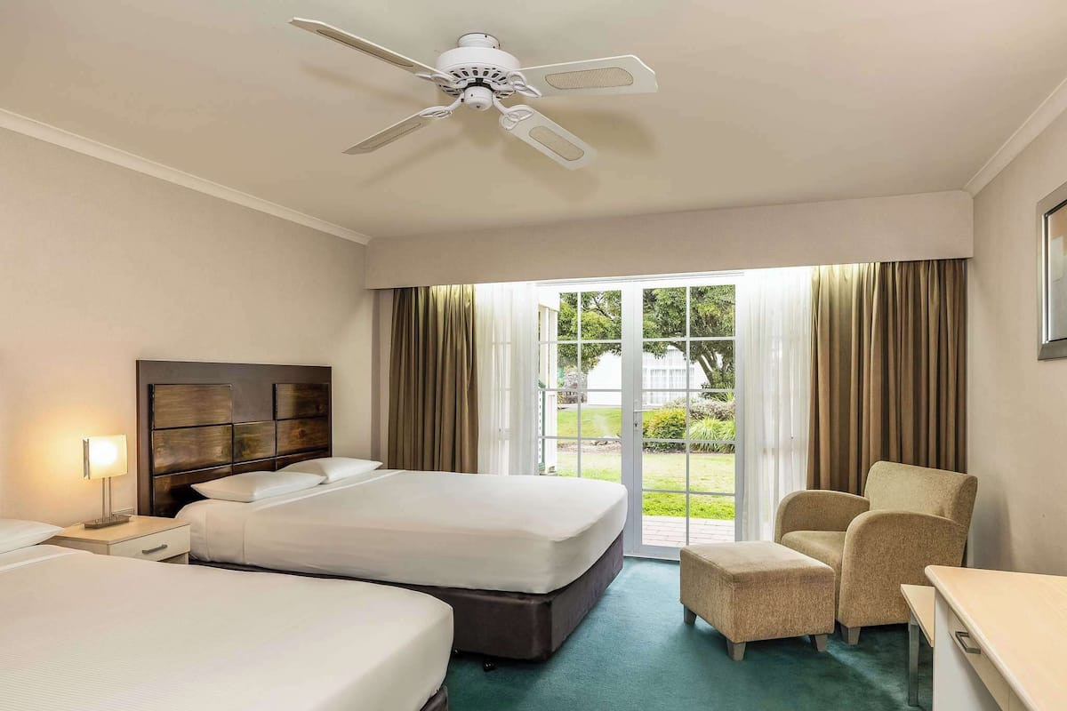 Habitación de hotel con dos camas, un escritorio, una silla, un ventilador de techo y vistas a través de puertas de vidrio.