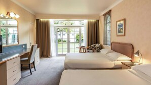 A hotel room with a desk, two beds, a chair, a small table, and a window with curtains.