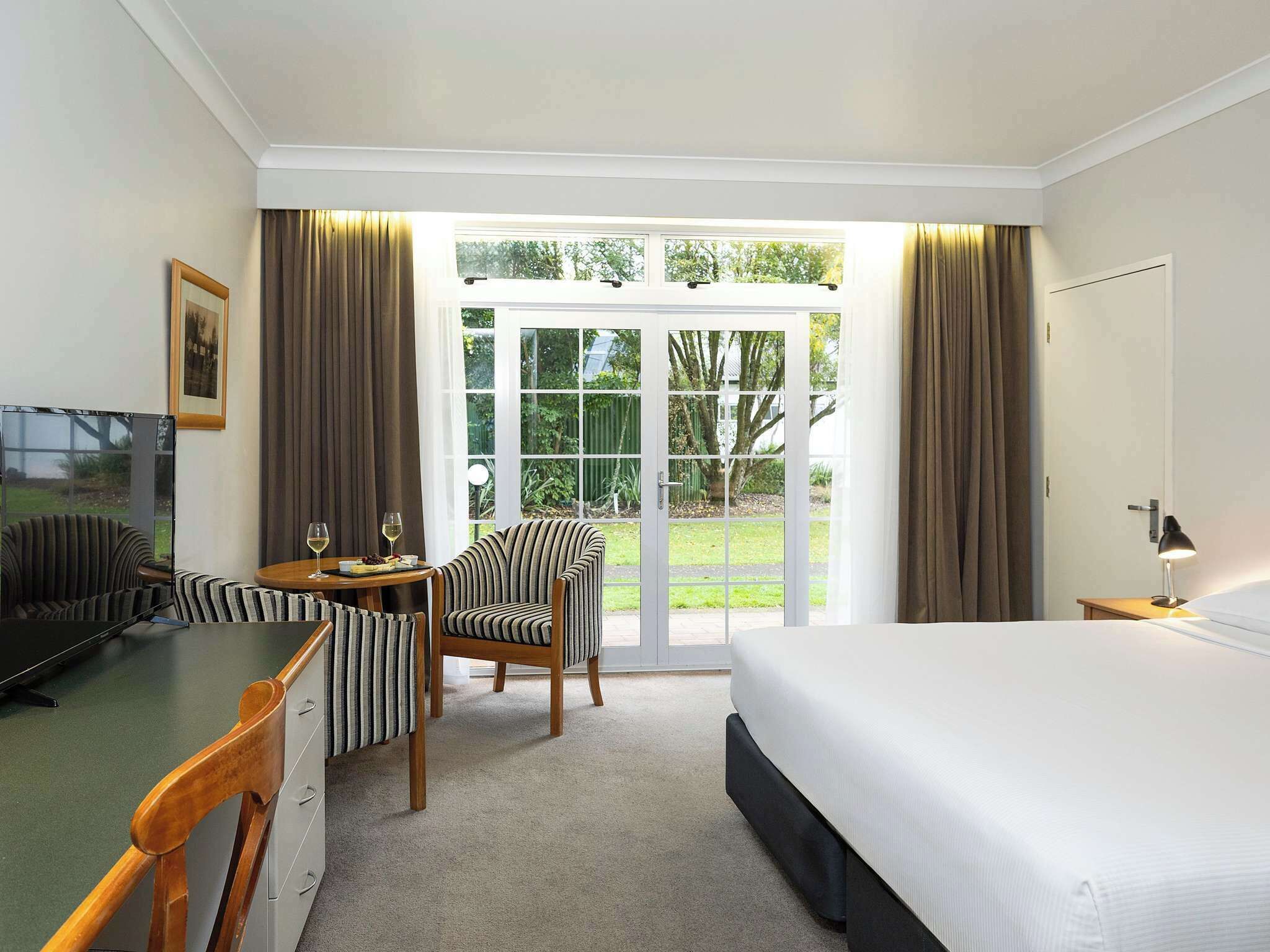 Ein Hotelzimmer mit einem Bett, einem Schreibtisch mit Stuhl, einem Fernseher und Blick ins Freie durch eine Glasdoor.