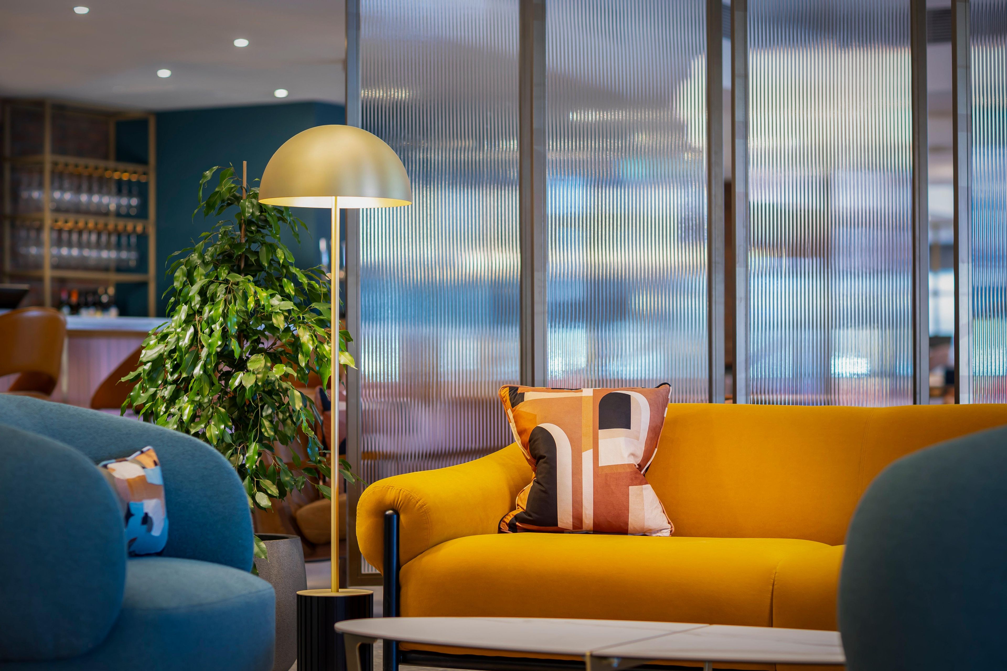 A modern interior with a yellow sofa, a blue armchair, a floor lamp, and a large plant.