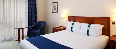 A hotel room with a large bed, a chair, a bedside table with a lamp, and a window with curtains.
