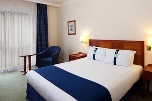 A hotel room with a large bed, a chair, a bedside table with a lamp, and a window with curtains.