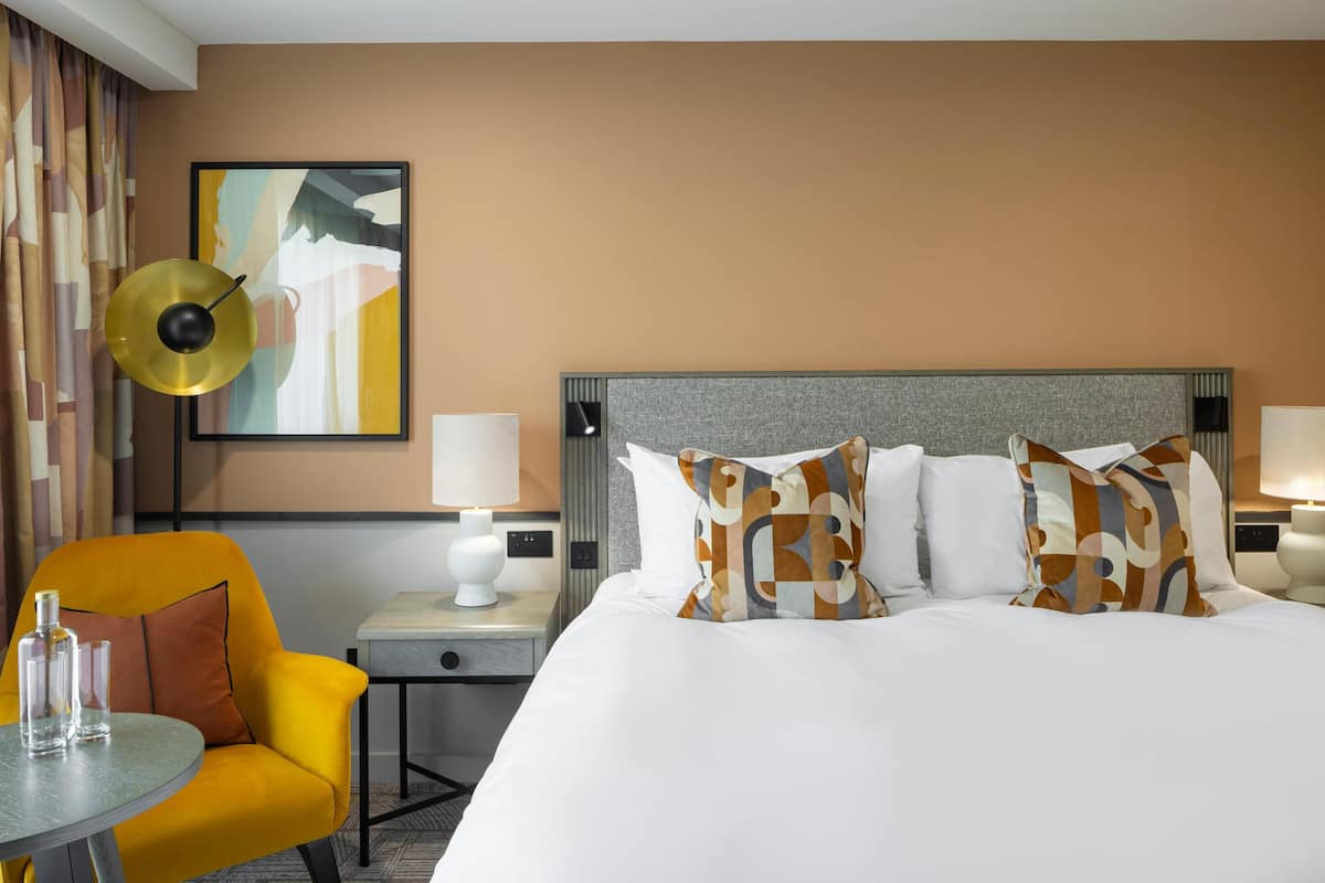 A hotel room with a bed, a chair, a table, a lamp, and a painting on the wall.