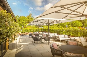 An outdoor patio area with tables, chairs, and umbrellas, surrounded by greenery and a brick wall.