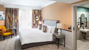 Habitación de hotel con cama, una silla, mesita de noche, lámpara, espejo y baño con lavabo y espejo.