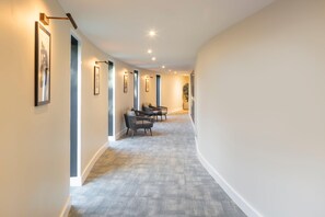 A hallway with framed pictures on the walls, a seating area with chairs, and a carpeted floor.