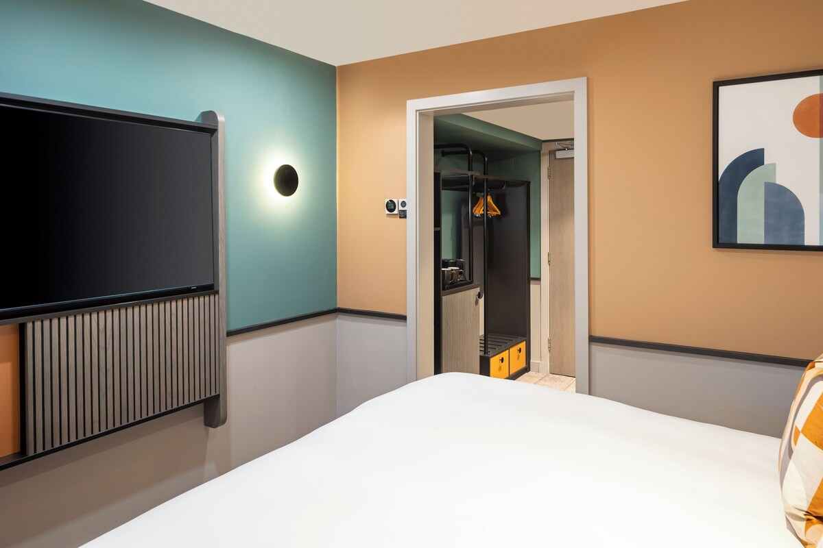 Una habitación de hotel con cama, televisor montado en la pared, radiador y puerta que da al baño.