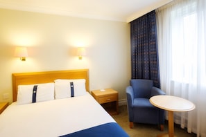 A hotel room with a bed, two bedside lamps, a chair, a small table, and a window with curtains. - Voco Fareham-solent (Fareham)