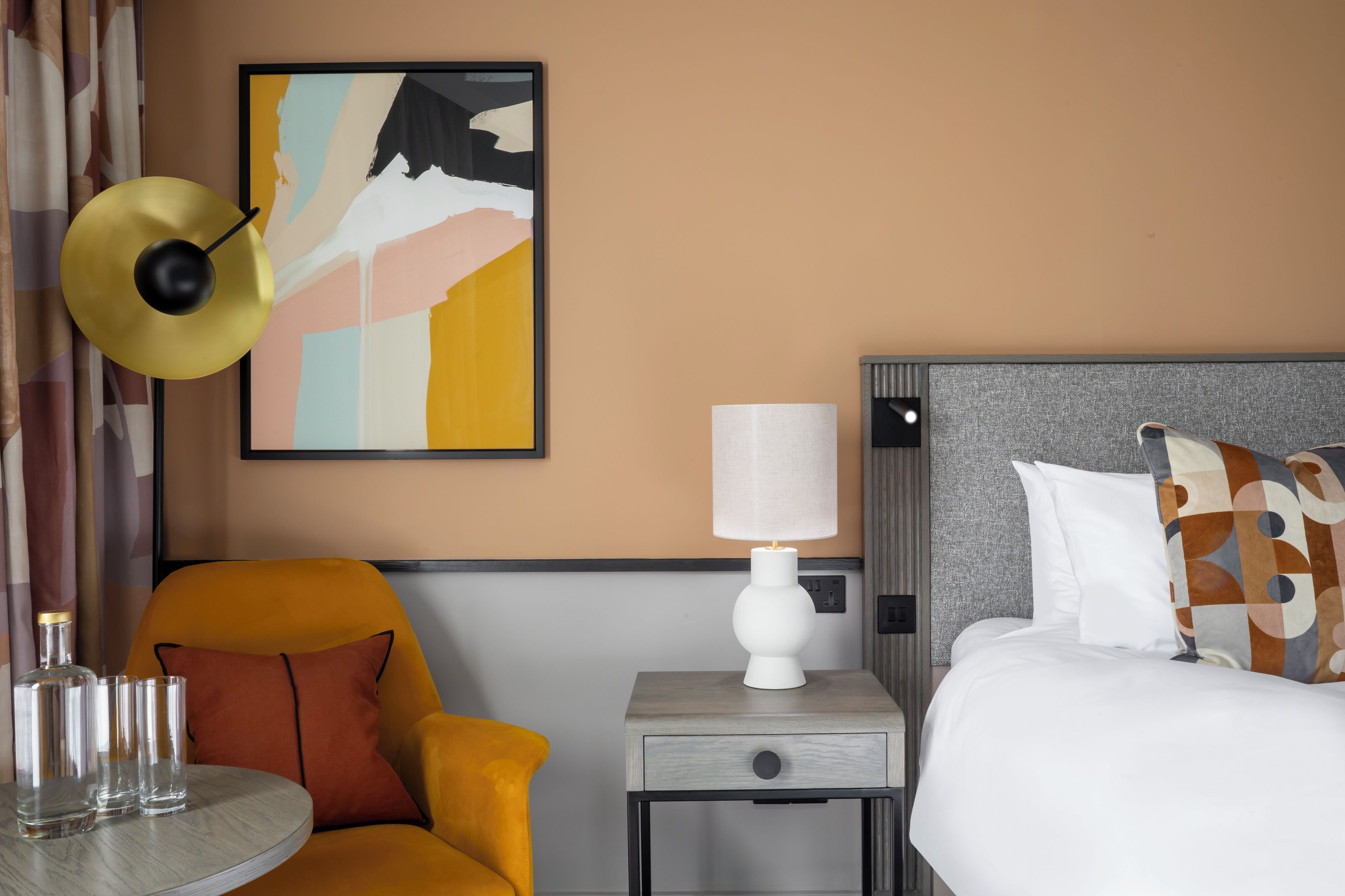 A hotel room with a bed, a nightstand with a lamp, a chair, a table with glasses, and a painting on the wall.