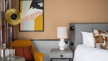 A hotel room with a bed, a nightstand with a lamp, a chair, a table with glasses, and a painting on the wall.