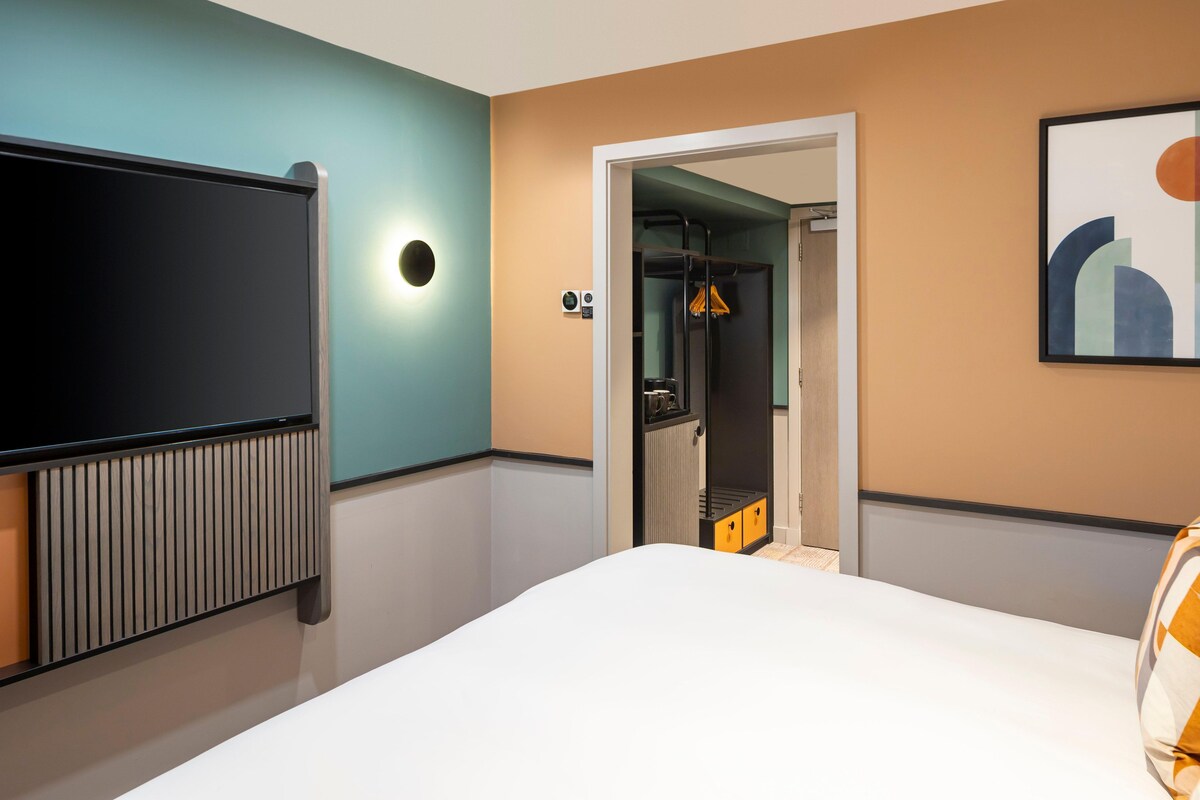 Habitación de hotel con cama, televisor, radiador, puerta que da a un baño y un cuadro en la pared.