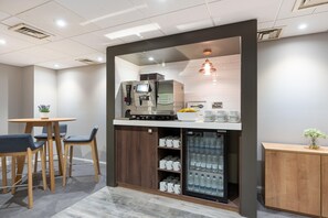 A compact hotel room with a kitchenette, a seating area with chairs, a small table, a refrigerator, and a coffee machine.