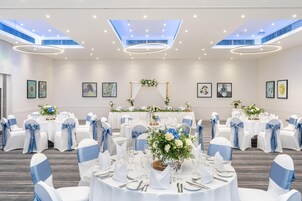 Un salón de banquetes con mesas redondas, sillas blancas y azules, e iluminación en el techo.
